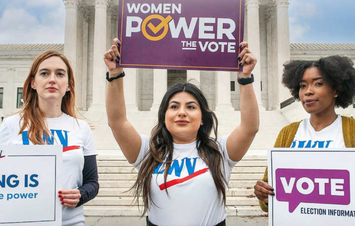 Three women (one white, one Latina, and one Black) hold voting signs outside the US Capitol building